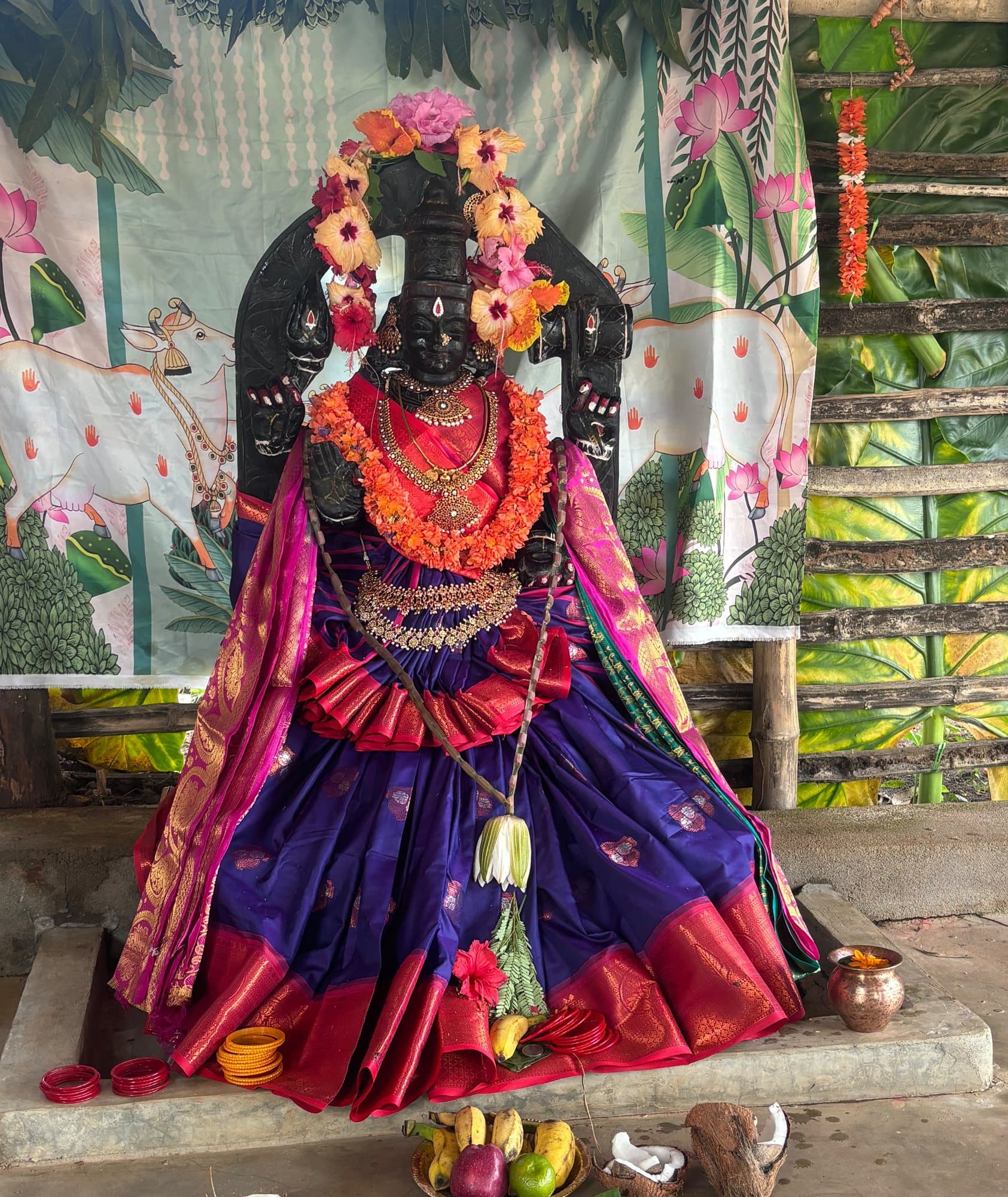 The murti of Maa Lalitha Devi in dark stone, adorned in a violet and red silk saree, gold jewellery and garlands of orange and pink flowers, before a painted backdrop of lotuses and cows.
